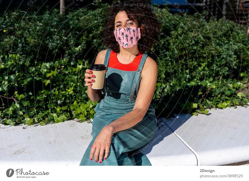 Geschützte Frau mit lockigem Haar sitzt an einem sonnigen Tag, schaut in die Kamera und trägt eine spezielle Maske Mundschutz Virus Krankheit Seuche Schutz