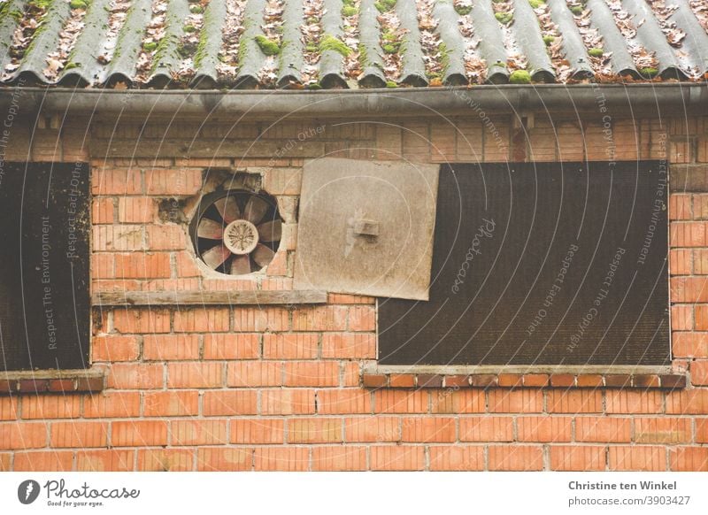 zeitgeschichte | Ventilator in der Außenwand eines alten landwirtschaftlichen Gebäudes mit Blechdach. Die Abdeckplatte ist zur Seite geschoben. Einige Steine beginnen abzubröckeln. Die daneben liegenden Fensteröffnungen sind mit Holzplatten verschlossen.