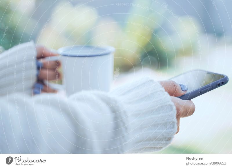 Nahaufnahme von Frauenhänden, die eine heiße Tasse Kaffee oder Tee halten und ein Smartphone im Hintergrund bei kaltem Wetter benutzen und warme, pelzbezogene Strickkleidung tragen