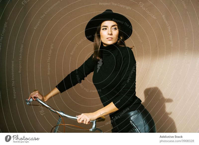Stilvolle Frau mit Hut steht in der Nähe von Fahrrad trendy Leder selbstbewusst Mode schlank jung brünett attraktiv schwarz Outfit Vorschein cool Kopfbedeckung