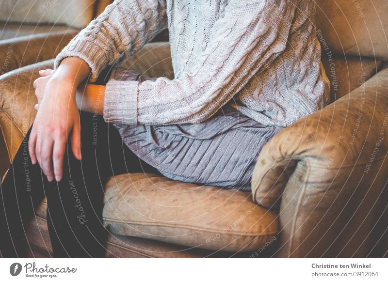 Eine junge Frau sitzt mit übereinander geschlagenen Beinen schräg in einem ausgeblichenen braunen Ledersessel. Sie trägt einen grauen Zopfpullover, einen grauen plissierten Rock und schwarze Strümpfe. Porträt ohne Kopf