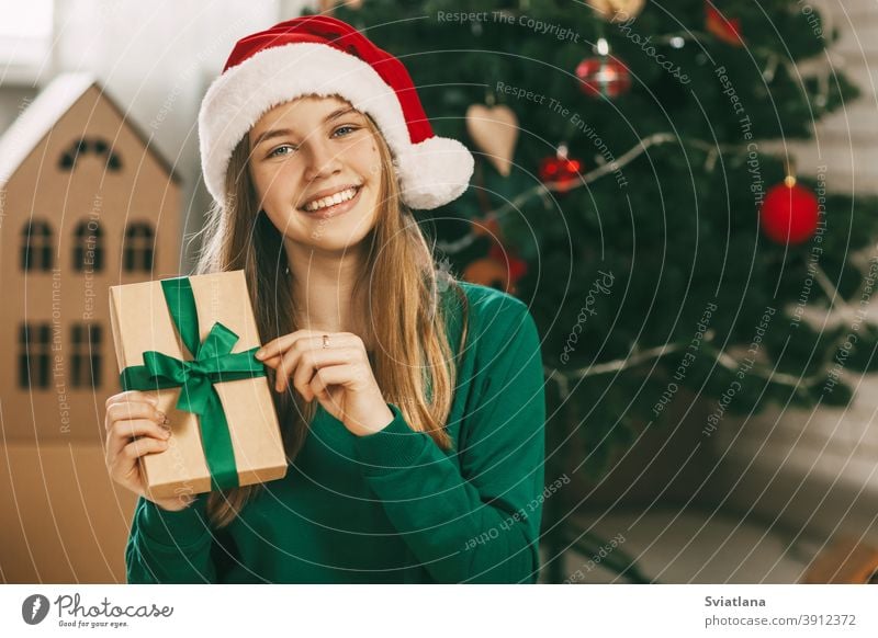 Ein hübsches kleines Mädchen schaut in die Kamera und lächelt. Sie hält ein Geschenk aus Kraftpapier, das mit einer grünen Schleife gebunden ist, in den Händen. Weihnachtsstimmung. Konzept für Neujahr Urlaub zu Hause.