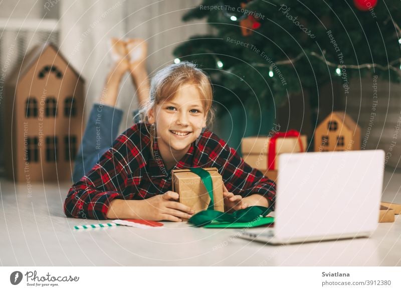Ein glückliches kleines Mädchen öffnet ein Geschenk unter dem Weihnachtsbaum zu Hause und kommuniziert mit ihren Großeltern über einen Laptop per Videoverbindung. Feiertagskonzept für Weihnachten und Neujahr zu Hause.
