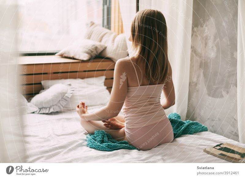 Eine junge Frau im Pyjama sitzt auf einem Bett in der Lotus-Position, die Finger in einer Mudra-Geste gefaltet, und genießt einen Morgen tiefer Achtsamkeitsmeditation. Rückansicht