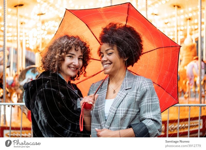 Fröhliche Freunde im Regentag Regenschirm Afro-Mädchen schwarze Frau Kaukasier Stadtleben Lächeln Vorderansicht Porträt Frauen in die Kamera schauen Karussell