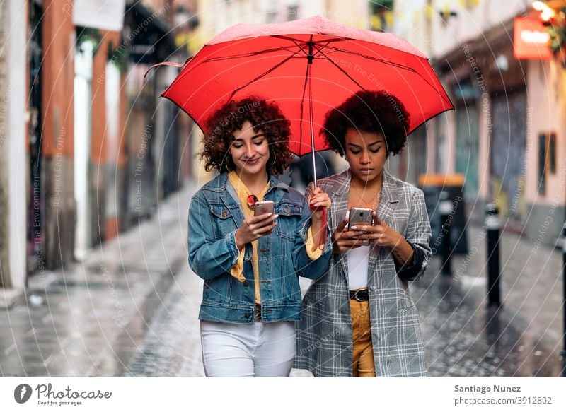 Glückliche Freunde mit Telefon Regenschirm Regentag Afro-Mädchen schwarze Frau Kaukasier per Telefon Stadtleben Lächeln Vorderansicht Porträt Frauen Blick