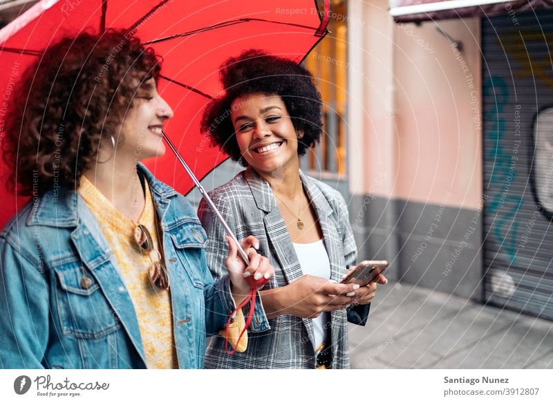 Fröhliche Freunde im Regentag Regenschirm Afro-Mädchen schwarze Frau Kaukasier per Telefon Stadtleben Lächeln Seitenansicht Porträt Frauen