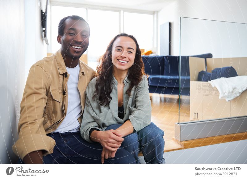 Porträt des jungen Paares, das in ein neues Haus umzieht und auf dem Boden im Wohnzimmer mit Umzugskartons sitzt junges Paar Hauskauf in die Kamera schauen