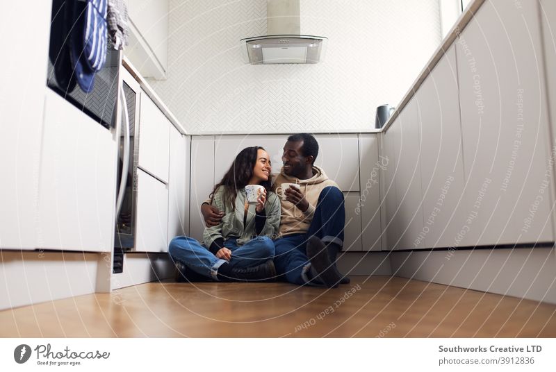 Junges Paar feiert den Umzug in ein neues Haus, das auf dem Boden mit einem heißen Getränk in der Küche sitzt junges Paar Hauskauf trinken Heißgetränk