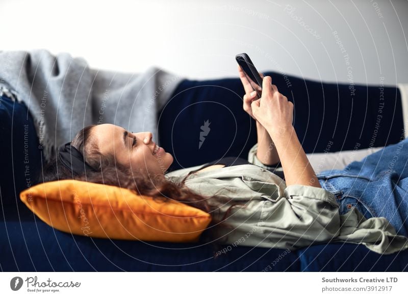Junge lächelnde Frau entspannt sich zu Hause auf dem Sofa liegend und überprüft soziale Medien auf dem Handy Lügen räkeln Mobile Zelle Smartphone Technologie