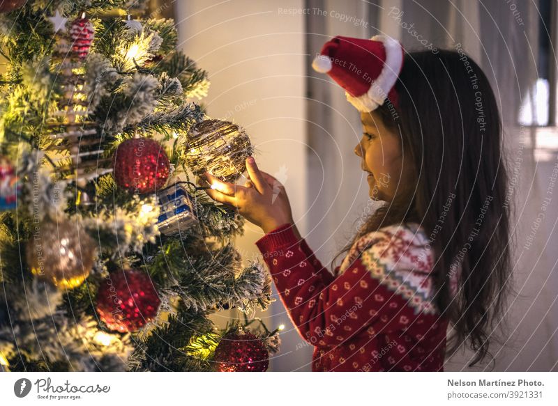 Kleines Mädchen in einem Weihnachtspulli und einer Weihnachtsmütze, die Weihnachtskugeln an den Baum hängen. Nizza offen schön Zauberei u. Magie Hintergrund