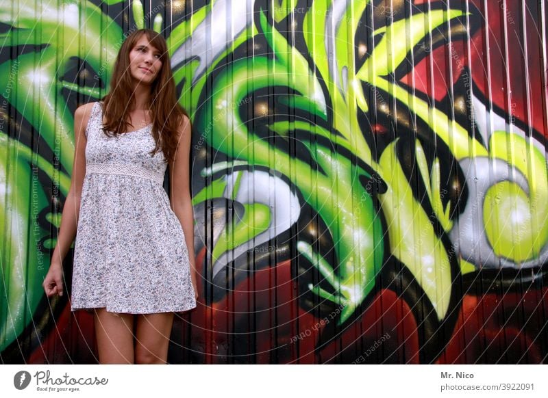 Junge Frau steht vor einer Grafitti Wand und schaut schüchtern nach links grafitti Sommerkleid Kunst Schriftzeichen Straßenkunst Fassade langhaarig Kultur grün