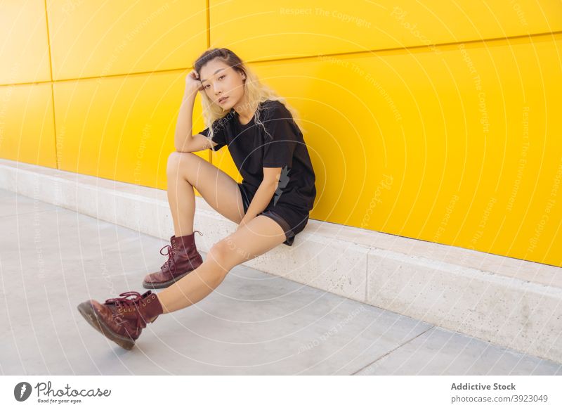 Junge ethnische Frau sitzt in der Nähe von gelben Wand Hipster jung urban Straße Stil Farbe Teenager modern asiatisch gefärbtes Haar tausendjährig Lifestyle