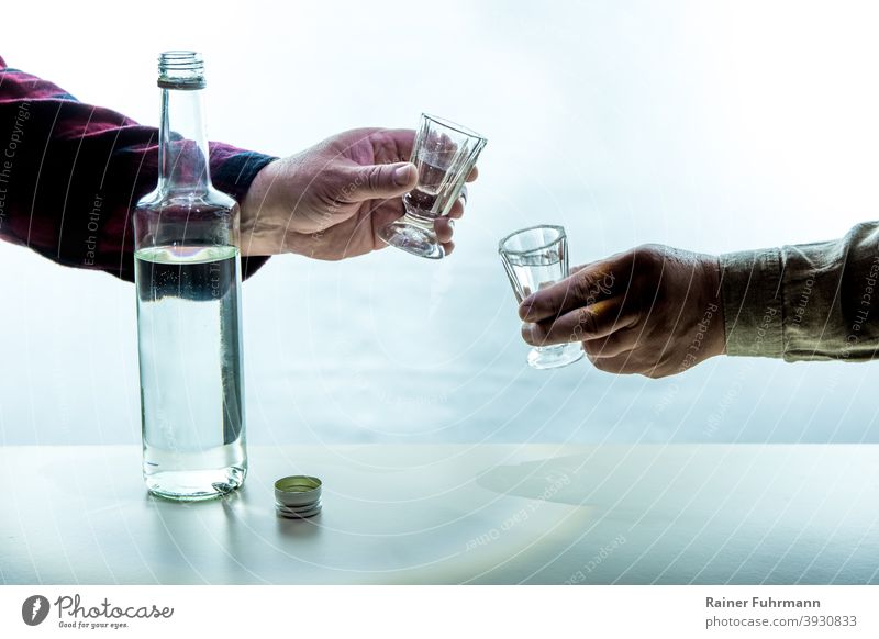 Auf einem Tisch steht eine Flasche mit Alkohol. Die Hände von zwei Männern halten Schnapsgläser. Sie stoßen miteinander an. Alkoholismus Sucht Feier feiern