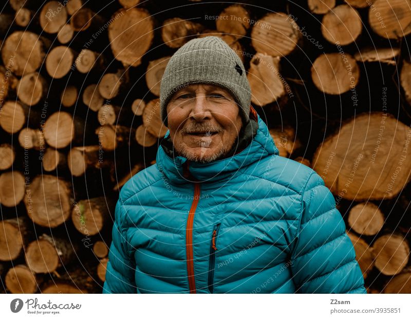 Porträt eines sportlichen Rentners vor einem Holzstapel spazieren Natur im Freien Sport rentner Ändern Mann mütze Winter Kälte Landschaft Wald sträucher Isar