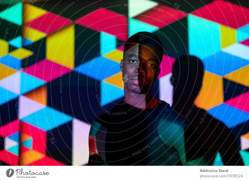 Ethnische Mann im dunklen Studio mit bunten Neon-Beleuchtung Atelier neonfarbig Licht Würfel Geometrie leuchten lebhaft farbenfroh Model männlich schwarz