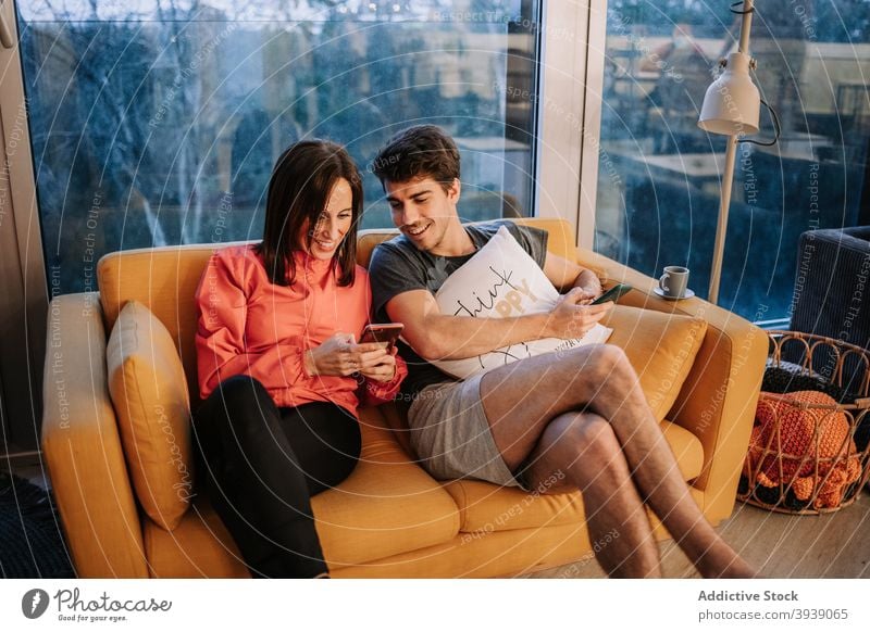 Lächelnde Freunde sitzen auf dem Sofa und verwenden Smartphone benutzend Zusammensein sich[Akk] entspannen heimwärts Wochenende zuschauen Video Mobile Telefon
