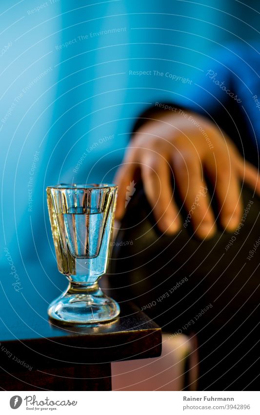 Auf einem Tisch steht ein altes Schnapsglas. Vor blauem Hintergrund sind Hand und Arm einer Person zu sehen. Alkohol Wodka Alkoholismus Alkoholiker Glas trinken
