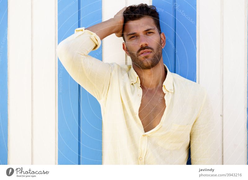 Junger Mann, Modemodell, posiert vor einer Strandbude. Frisur Sommer modisch Vollbart Model Blick Person Typ jung Hemd gutaussehend Porträt Hintergrund