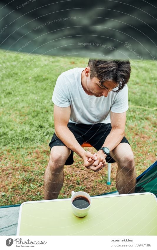 Junger Mann trinkt einen Kaffee sitzend vor dem Zelt am Morgen Abenteuer Getränk Pause Frühstück Lager Camping Nahaufnahme Tasse trinken Erkundung Freiheit