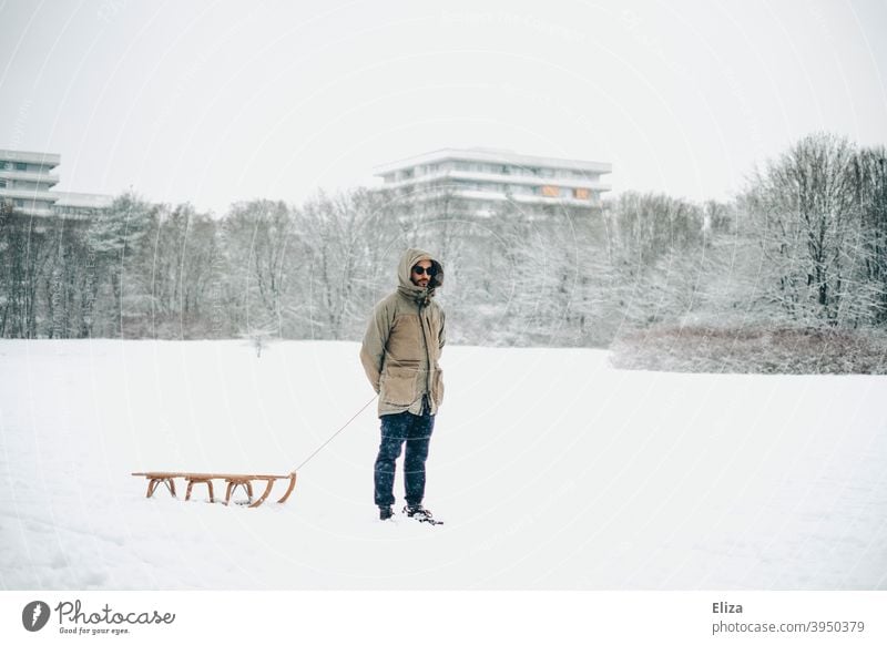 Mann mit Schlitten im Schnee und Hochhäusern im Hintergrund Wiese Natur Mensch Schlittenfahren Holzschlitten Winter Wintertag grau weiß kalt Schneelandschaft