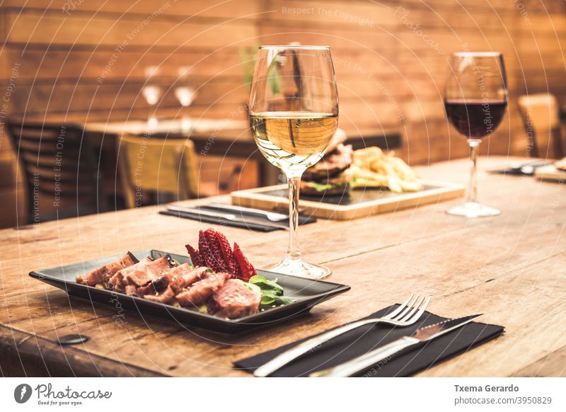 Traditionelle Restaurantatmosphäre mit Thunfisch-Tataki und Burger mit Pommes im Hintergrund auf rustikalem Holztisch und einem Glas Weißwein Fisch Chips