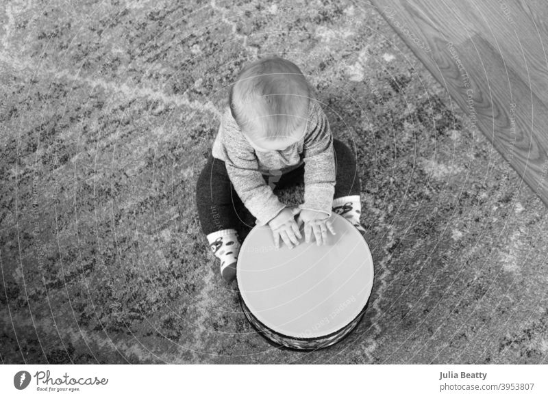 6 Monate altes Baby spielt Trommel, während es auf einem Teppich auf dem Boden sitzt; Schwarz-Weiß-Bild aus der Vogelperspektive Bodentrommel Hände schlagen