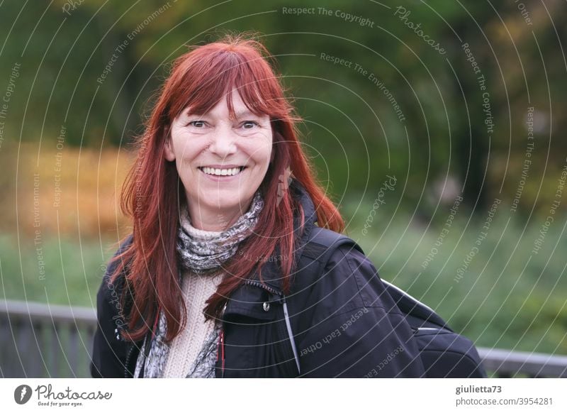 Glückliche, lächelnde Frau mit roten langen Haaren draussen in der Natur Blick in die Kamera Vorderansicht Oberkörper Porträt Zentralperspektive