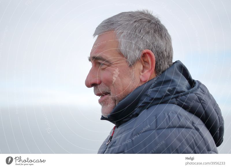 Porträt eines lächelnden Senioren im Profil mit grauen kurzen Haaren, Dreitagebart und blauer Winterjacke mit Kapuze Mensch Mann männlich kurzhaarig grauhaarig