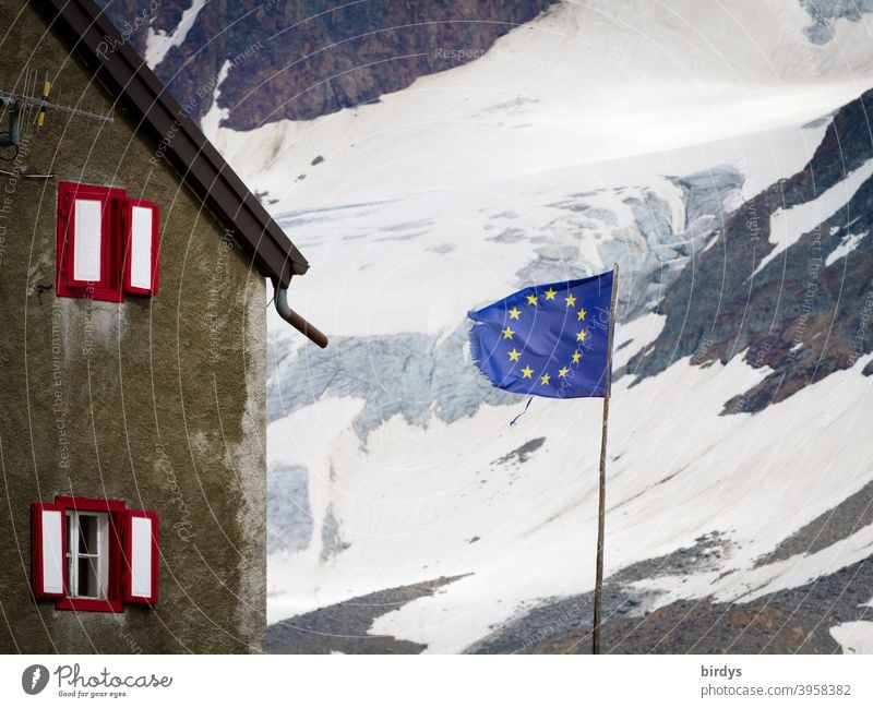 Flagge der europäischen Union vor einem Haus in den Bergen , im Hintergrund ein schneebedeckter Berghang . EU - Fahne Europäische Union Europafahne Alpen Schnee