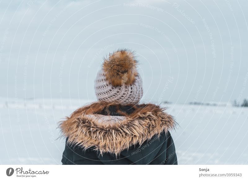 Rückansicht von junger Frau mit Wollmütze und Jacke im Schnee Winter Außenaufnahme kalt Einsamkeit friedlich betrachtend weiß Pelzkragen Farbfoto Tag Frost