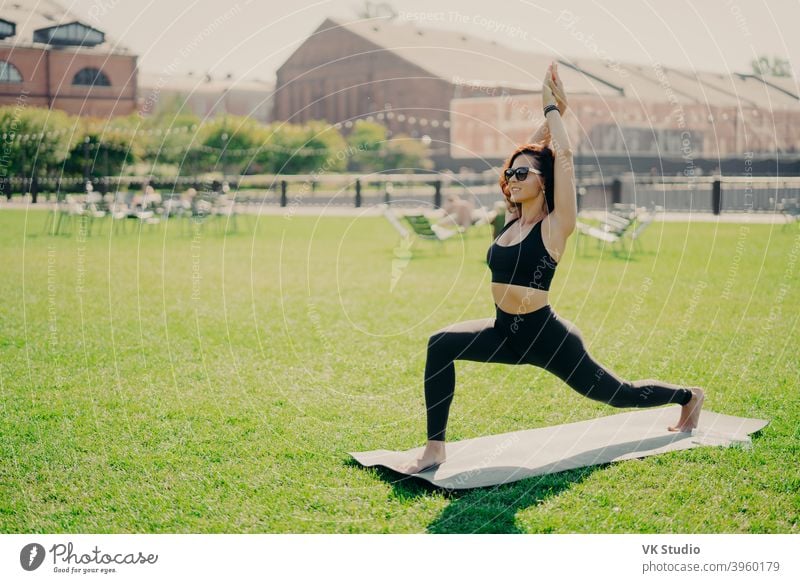 Konzentrierte positive Frau hebt Arme tut Pilates-Übungen in aktive Kleidung Sonnenbrille gekleidet hat Training im Sommer Praktiken Yoga-Posen auf grünem Gras. Menschen und gesunden Lebensstil Konzept