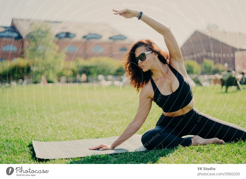 Bild von motivierten fit Frau hebt Arm und macht Dehnübungen im Freien posiert auf Fitness-Matte trägt Sonnenbrille und activewear Züge aktiv. Fitness-Trainer geht in für Sport im Freien