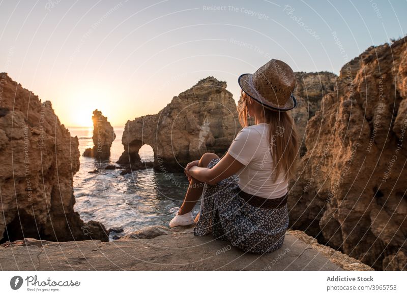 Unerkennbare Frau rastet in der Nähe von Meer und Klippen ruhen MEER bewundern Sonnenuntergang Wasser Natur Küste Stein Algarve Portugal Urlaub sitzen Hut