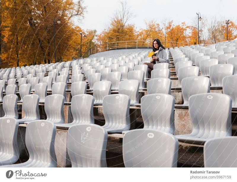 junges Mädchen im grauen Mantel sitzt auf einem Sitz in einem leeren Stadion Frau Sitzen Herbst allein Traurigkeit einsam Kaukasier schön Emotion im Freien