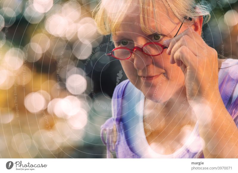 AST10  l Eine sympathische Frau schaut über ihre Brille, hält sie fest und lächelt leicht. Sie ist umrahmt mit vielen Lens -Lichtern Lächeln freundlich