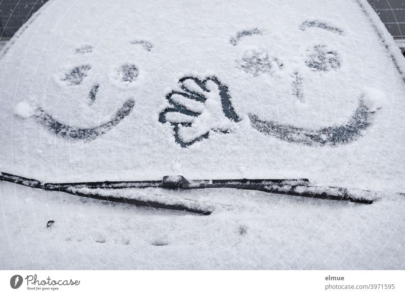zwei Gesichter und eine Hand sind auf der Frontscheibe eines verschneiten Autos zu sehen / Winterspaß Schnee Scheibenwischer malen freikratzen zugeschneit weiß
