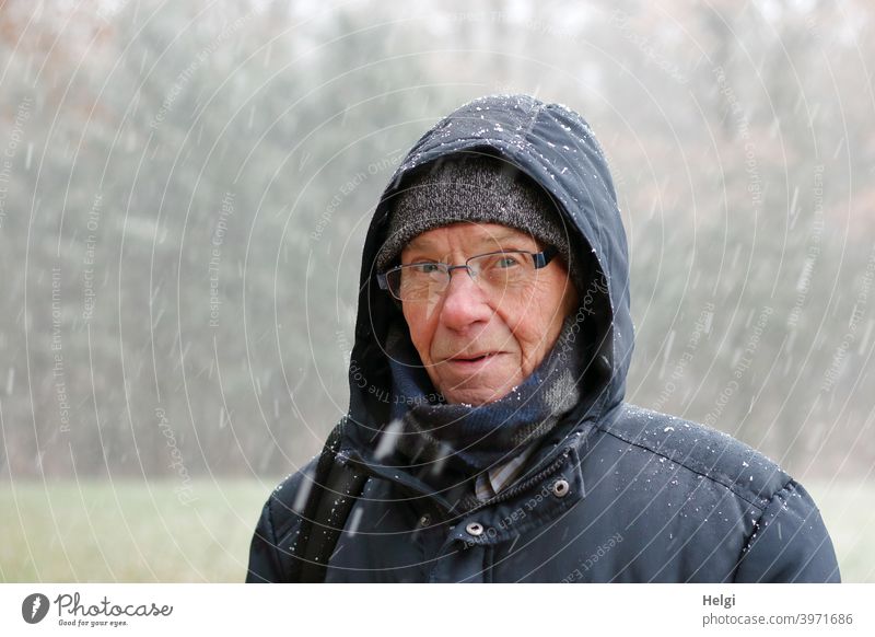 Porträt eines Seniors, der mit Winterjacke, Schal, Mütze und Kapuze im Schneegestöber steht Mensch Mann Gesicht Brille Schneefall Kälte Blick frieren draußen