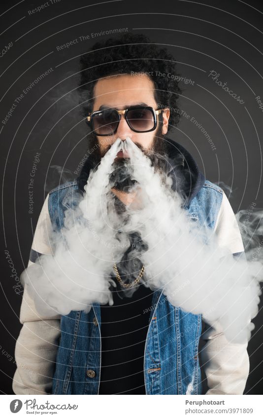 Junger Mann vaping, Studioaufnahme. Bärtiger Mann mit Sonnenbrille bläst eine Rauchwolke auf schwarzem Hintergrund. Konzept des Rauchens und Dampf ohne Nikotin.