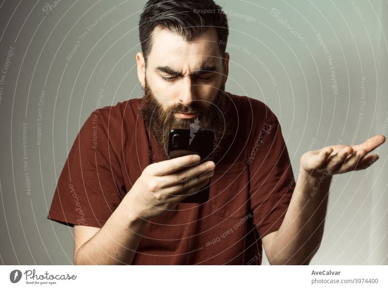 Ein junger Mann im Hipster-Stil mit Bart und modernem Haarschnitt wütend, während er auf seinen Telefonbildschirm mit Kopierraum schaut Angst verwirrt Fehler