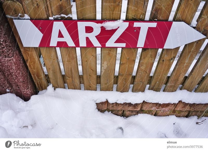 Schild als großer Pfeil und mit der Aufschrift "ARZT" in Druckbuchstaben ist an einem Holzlattenzaun befestigt / ärztliche Hilfe / Orientierung Arzt Hinweis