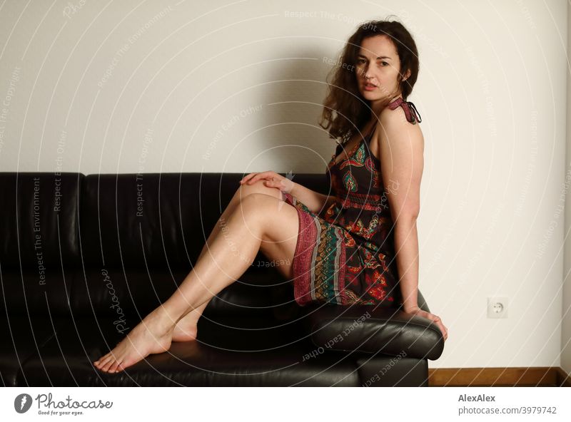Porträt einer jungen, brünetten, langhaarigen Frau, die barfuß auf einer dunklen Couch im Sommerkleid sitzt natürlich hübsch lange Haare schlank Schulter Haut