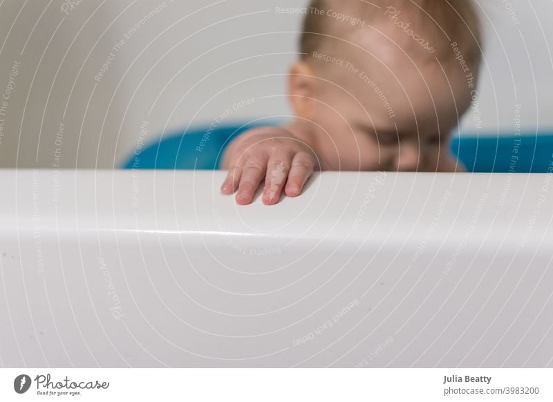 Nahaufnahme von Händen und Fingern des Babys, die den Rand der Badewanne greifen; im Hintergrund sitzt das Baby aufrecht in der blauen Babybadewanne Badezeit