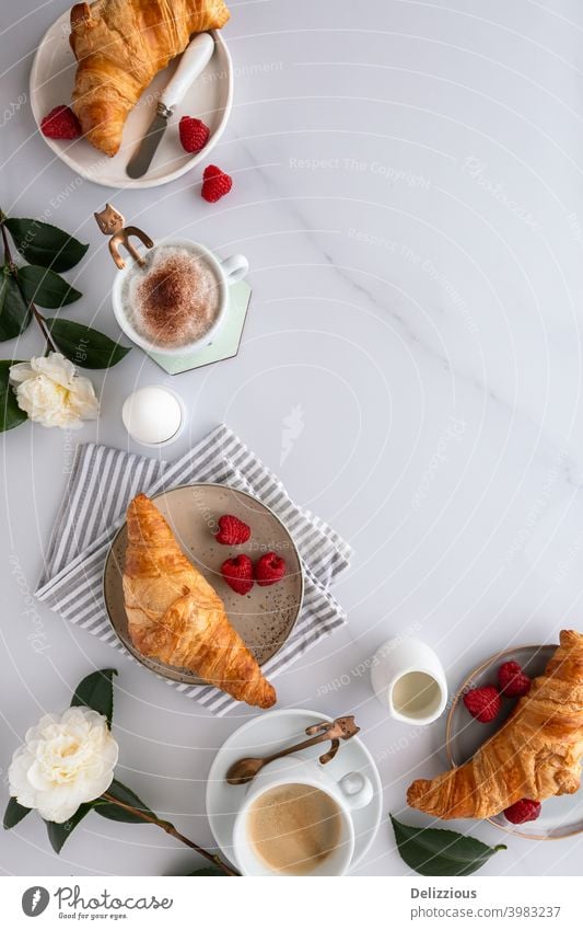 Blick von oben auf einen Frühstückstisch mit Croissants, Kaffee, Himbeeren, Blutorange, gekochtem Ei und Blumen auf einem weißen Holztisch, auf weißer Marmorfläche