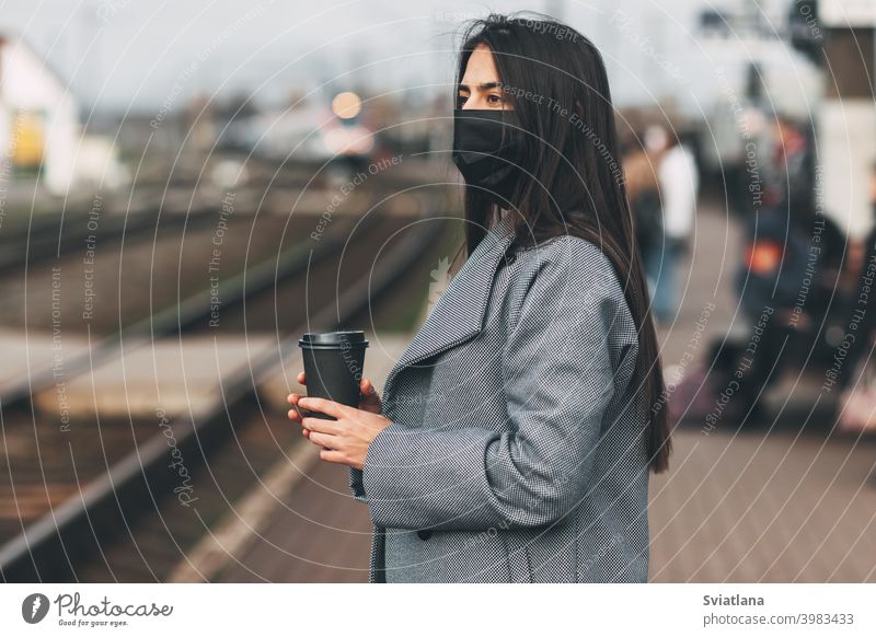 Fröhliches Mädchen in einer Maske steht mit Kaffee in der Hand am Bahnhof und wartet auf den Zug. Verkehr jung Frau schön Eisenbahn Passagier U-Bahn Lächeln