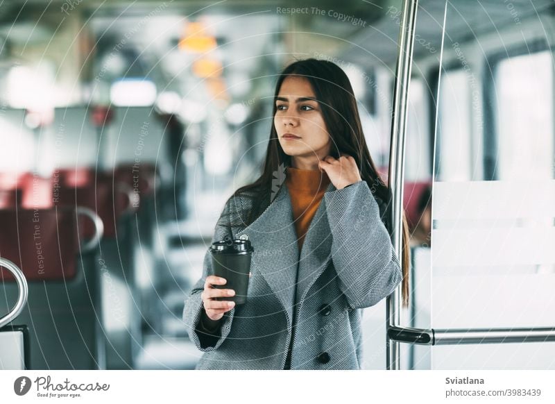 Eine fröhliche Brünette mit Kaffee in der Hand steht im Zug, schaut in die Ferne und lächelt. Verkehr jung Frau schön Eisenbahn Passagier U-Bahn Lächeln