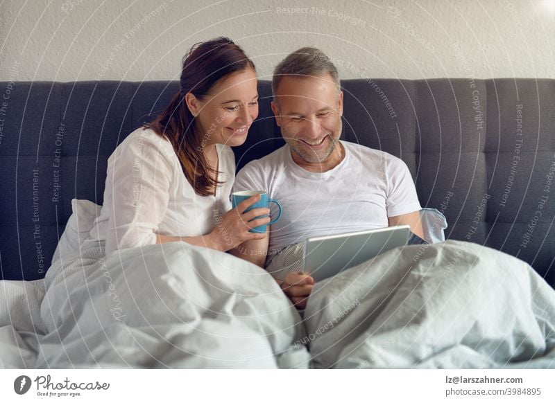 Glückliches Paar mittleren Alters, das morgens im Bett sitzt, Kaffee trinkt und mit einem digitalen Tablet Online-Einkäufe tätigt Sofa Möbel Familie Raum