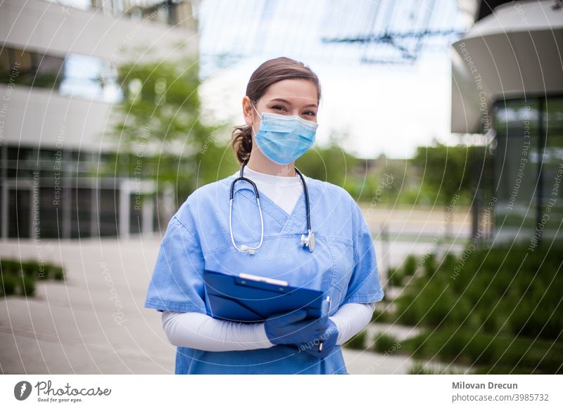 Arzt vor einem Krankenhaus oder Pflegeheim, in blauem Kittel, Gesichtsmaske, hält Klemmbrett mit Patientenformular betreutes Wohnen Gebäude Rekonvaleszent