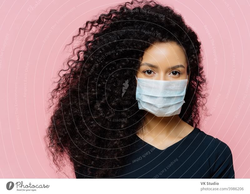 Ernste dunkelhäutige junge Frau, die zu Hause auf Selbstisolierung ist, trägt eine medizinische Schutzmaske, ist zu Hause auf Quarantäne, ist leger gekleidet, posiert vor rosa Hintergrund. Coronavirus-Krankheit