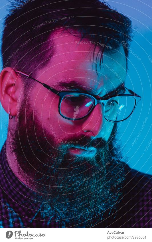 Ein junger Hipster-Mann mit blauer Lichtbrille schaut mit ernstem Gesicht auf blauen und rosa Farben zur Kamera weg 20s Kontemplation Unternehmer genial
